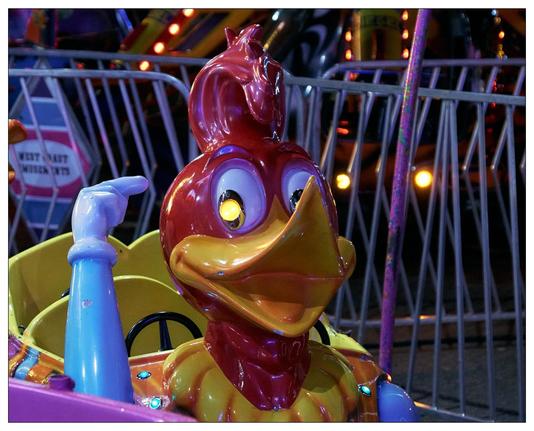 The image shows a colorful ride featuring a cartoonish, bright red and red woodpecker. This is part of a children's amusement park ride, indicated by its whimsical design and the safety bars visible in the background. The ride element appears to be a small car or carriage, designed for young children to sit in during the ride. The background includes dimly lit amusement park lights and fencing that typically surrounds such rides for safety.