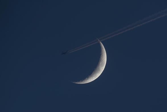 Der zunehmende Mond am dunkelblauen Dämmerungshimmel mit einem Flugzeug mit Kondensstreifen die Sichel oben querend