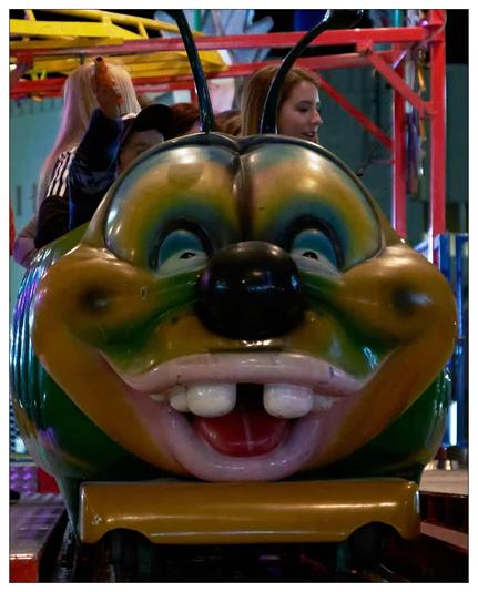 This is an image of a colorful amusement park ride designed to look like a caterpillar's face, featuring exaggerated facial features like big eyes and a wide smile. There are four individuals, possibly enjoying a ride inside this caterpillar-shaped cart. The background shows parts of the ride structure and other equipment commonly found in indoor amusement areas. The image captures a moment of leisure and entertainment in a lively and colorful setting.