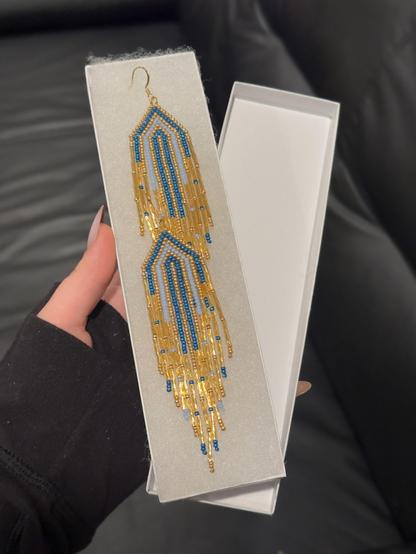 A hand in a black sleeve holding a pair of gold and blue beaded fringe style earrings in a white box against a black background