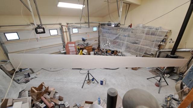 One glider wing sits diagonally in a garage. It is freshly painted white. It is positioned leading edge up. Various tools and clutter are in the corners of the garage, and wall shelves are covered in plastic drop cloth.