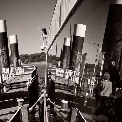 Image im B&W and voluntarily slightly inclined. To the left one sees the dock, to the right (occupying 2/3s of the image) the ship of which the windows reflect the dock and where 2 people disembarking seen from the back.