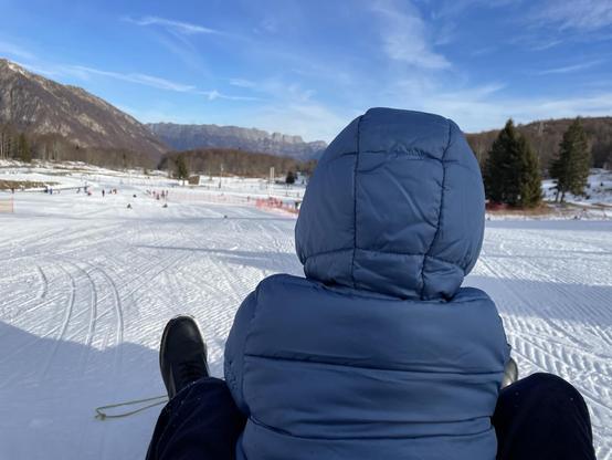 La foto mostra la schiena di un bambino seduto su uno slittino, con indosso un cappellino con cappuccio blu. Il bambino guarda verso una collina innevata con altre persone che slittano in lontananza. Sullo sfondo si vedono montagne innevate e un cielo azzurro.