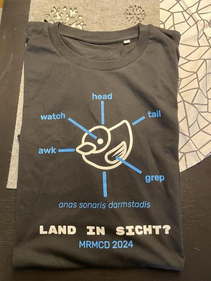 Ein schwarzes T-Shirt mit einer stilisierten Vogelillustration in weiß mit den Begriffen "watch", "head", "tail", "awk" und "grep" an verschiedenen Körperteilen in blau beschriftet. Darunter steht
anas sonaris darmstadis
LAND IN SICHT?
MRMCD 2024