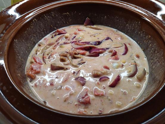 A slow cooker bowl made of brown china, filled with a sort of pinkish opaque sauce and rings of red onion floating therein.