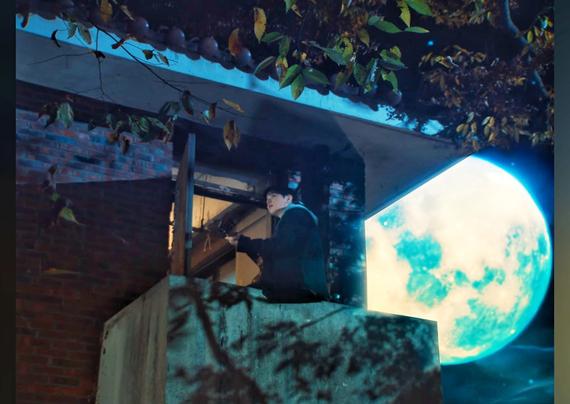 Seungmin playing guitar on a balcony with a huge blue moon in the background