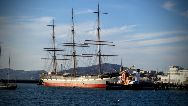 Ein Dreimaster liegt im Hafen ohne Segel. Der Rumpf ist längst in roten, grauen, weißen und schwarzen Streifen gestrichen. Davor liegt eine kleines Dampfschiff, dass ggf. eine Schlepper gewesen sein könnte. Das Meer ist Blau. Der Himmel fast wolkenlos.
