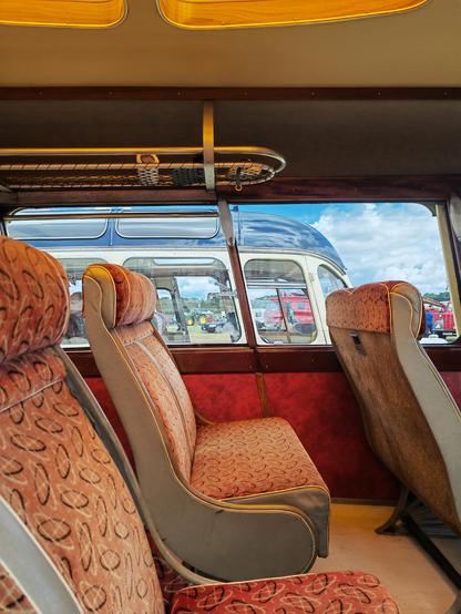 An interior view of a touring coach. The seat upholstery is orange-brown and the panelling scarlet. Dark stained wood and leather panels frame the brighter colours. The floor is a beige linoleum. The length of the coach is illuminated by skylights that are lined with a fruitwood or maple veneer with brass trim and the lights have an orange filter, casting a warm, sunny glow throughout. Behind, a coach of the same type in blue livery shows the arrangement of the skylights.