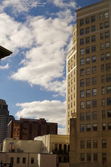 A view of a city skyline, featuring a mix of modern and older buildings. A tall, light brownish-tan brick building, likely a commercial or office building, dominates the right side of the image. In the foreground, a section of a light beige/tan building is visible, with a small balcony or rooftop terrace. Other buildings, including one with the "Comerica" logo, are partially visible in the mid-ground. The sky is partly cloudy, with a mix of blue and white clouds. The image suggests a sunny day, with shadows cast by the buildings.