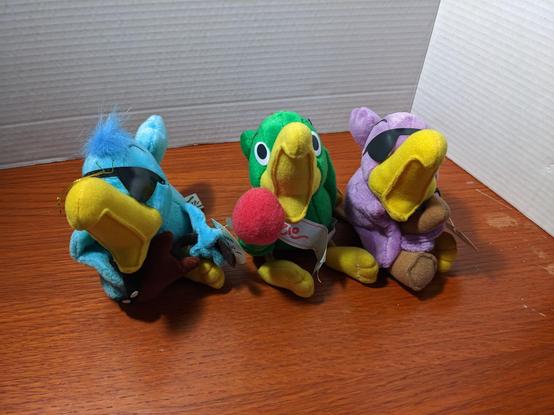 Three parrots, all yellow beaks, all wide open as if singing. The green parrot holds a red topped microphone, the blue parrot holds a brown guitar, the purple parrot holds a saxophone looking instrument.