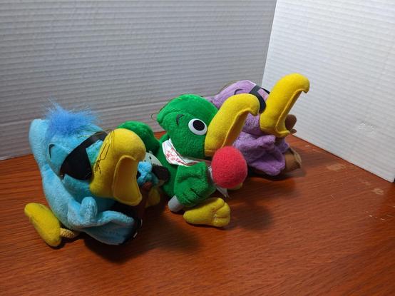 Three parrots, all yellow beaks, all wide open as if singing. The green parrot holds a red topped microphone, the blue parrot holds a brown guitar, the purple parrot holds a saxophone looking instrument.