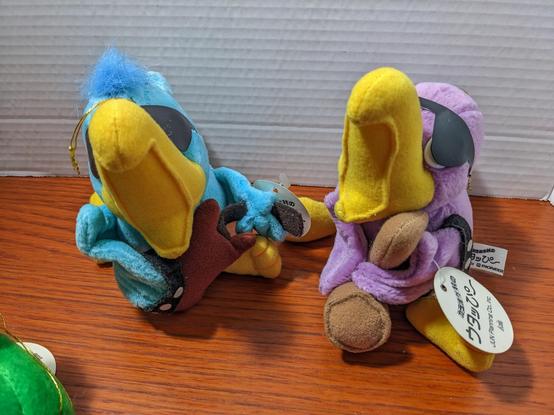 Two parrots, all yellow beaks, all wide open as if singing. The blue parrot holds a brown guitar, the purple parrot holds a saxophone looking instrument.