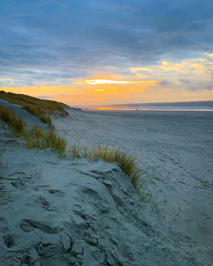 Blick über den Strand von Langeoog auf den Sonnenuntergang. Das linke Drittel des Bildes wird von der ansteigenden bewachsenen Düne beherrscht. Rechts im Hintergrund ist die Brandung der Nordsee zu sehen.