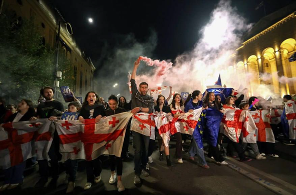 This picture shows a street in Georgia, in the darkness of the night, a wall of people demonstrating for freedom, holding georgian and european flags in front of their breasts. In the middle one holding a smoke grenade over his head, on the side the illuminated columms of a historic building.

the picture is remindful of  paintings of the french revolution. A composition of light and darkness.