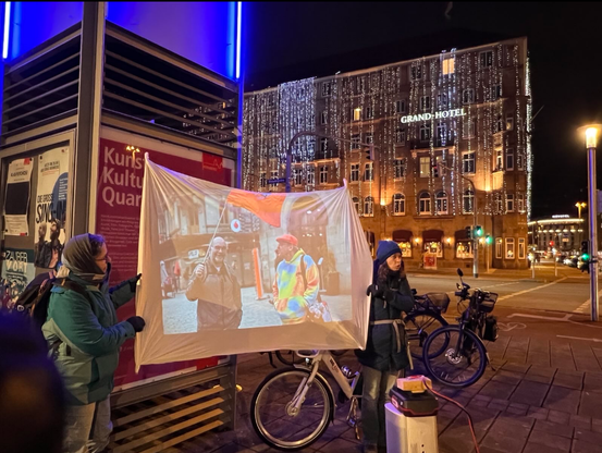 2 Menschen halten ein Leintuch gegenüber des Nürnberger Grand Hotel, das im Dunkeln mit leuchtenden "Eiszapfen"-Ketten dekoriert ist. Auf dem Tuch ist eine projizierte Szene mit 2 Aktivisti mit XR-Fahne zu sehen.