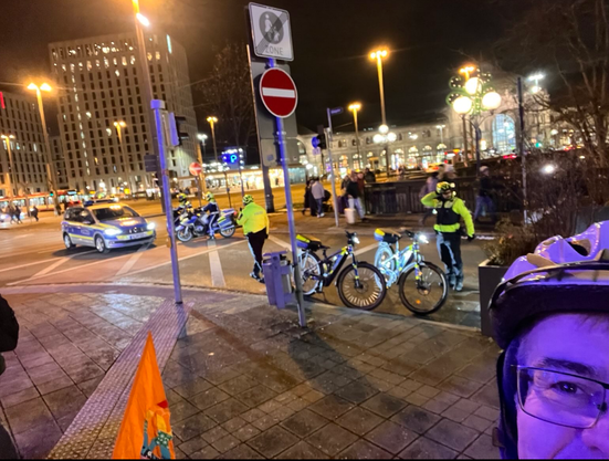 Polizei sichert eine mit diagonaler Gittermarkierung provisorisch eingerichtete Einmündung, die eine Fußgängerzone ist, mit einem Auto und zwei Polizeimotorrädern. Vorne dran stehen zwei Polizei-Fahrräder am Straßenrand. Es ist dunkel und neben den Straßenlampen ist im HIntergrund der Nürnberger Hauptbahnhof zu sehen.