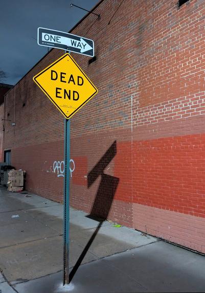 A ONE WAY/DEAD END Sign casts a shadow on the sidewalk and against a brick wall in the evening.
