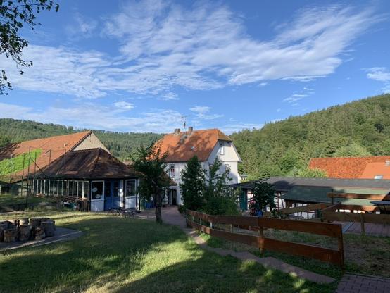 Foto des Geländes und der Gebäude des Schullandheims Lützels. Im Bild mehrere Gebäude, im Vordergrund Wiese, im Hintergrund bewaldete Hügel und blauer Himmel mit weißen Wolken.