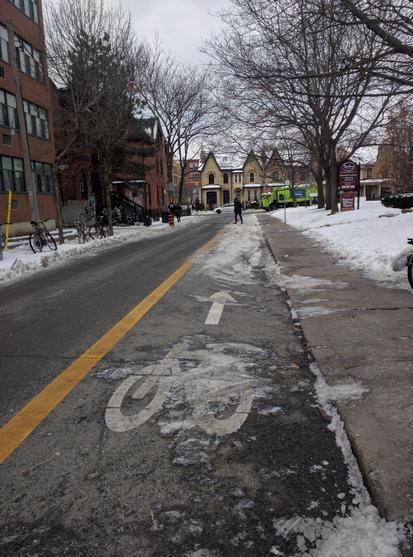 Bicycle lane with barely-visible markings, and an arrow pointing directly towards a pile of snow, covering the bike lane.