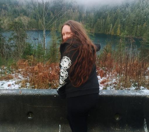 A person with long auburn hair wearing a black hooded sweatshirt with a moon phase pattern on the sleeve smiles at the camera while leaning on a concrete barrier. Behind them is a lake and a snowy forest.