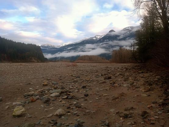 A dry riverbed with a scattering of rocks and stones. In the background, a mountain range stretches across the horizon, partially obscured by low-hanging clouds. The sky above is a mix of blue and white, with the sun casting a warm glow on the scene.