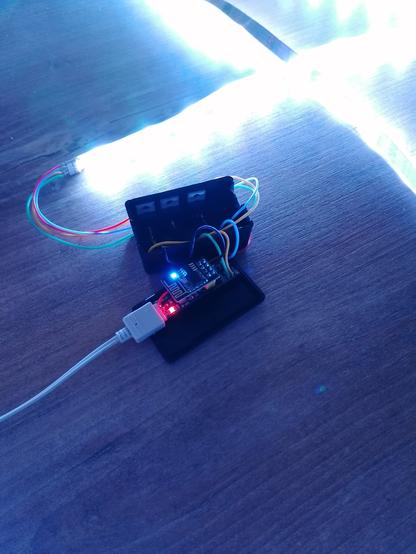 A open, black case holding electronics. 
In focus, a ESP-01 is mounted on a breakout bord. From left, a white cable is connected into the box, where a red light is indicating power and a blue light on ESP-01 is indicating action.
Wires are leaving the box to the right, where they are connedted to a bright shining LED stripe.