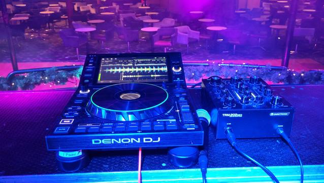 A Denon SC 6000 Prime media player, you might know with the big jog wheel and large touch screen display and a Omnitronic TRM-202 Mark 3 on a table on a stage. Between them a microphone. Also in the picture hues of magenta, a narrow strip of the dancefloor and tables and chairs.