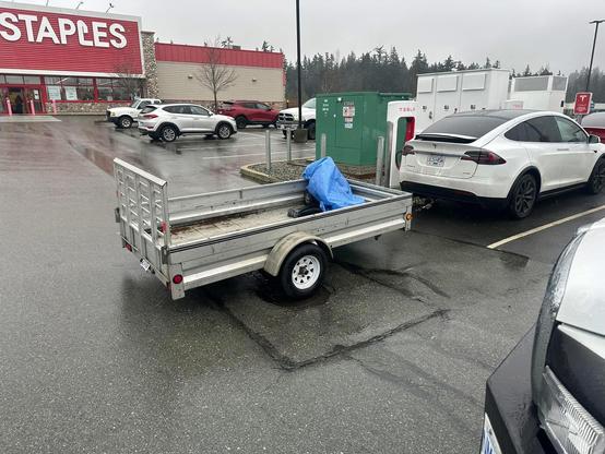 A small trailer is attached to a white Tesla. It is in a parking lot with a large staple store in the background. In the extreme foreground in the right corner is the front end of what looks like a large truck. Probably owned by the person who took the picture incredulously and posted it on the Facebook group.