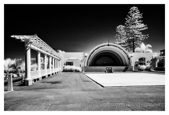 A black and white infrared photo of a small performance stage with a half dome roof and a tarmac and concrete apron in front. On the left is a colonnade and the architecture is in Art Deco style.