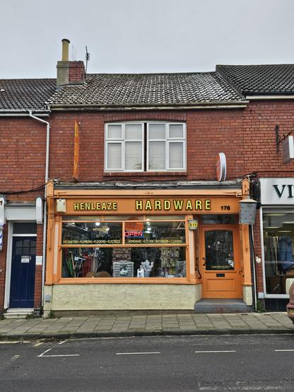 Photo of the shop front of 'Henleaze Hardware'. Orange door and frane with yellow san serif allcaps lettering outlined in black.