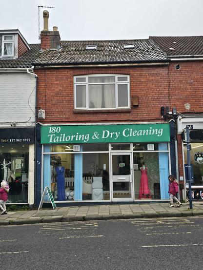 Photo of the shop front of 'Tailoring and dry cleaning'. Aluminium door with green sign background a white mixed case, serif with dtroke contast  lettering.