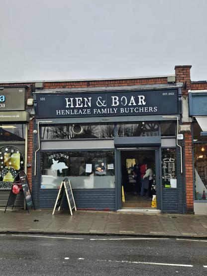 Photo of the shop front of 'Hen & Boar' butchers shop. Dark, slate blue door frontage with white distressed serif allcaps lettering shop name and the phrase 'Henleaze family butchers' below in a serif allcaps cobtrast with thinner stoke.