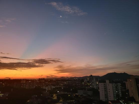 Sunset over a city with the moon and venues visible above and mountains in the background.