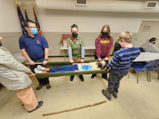 Three scouts and two leaders holding up another scout in a handmade stretcher. The stretcher was made using two scout staffs and a piece of canvas.