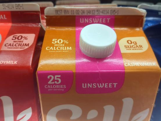 A photograph of the non-dairy milk section at the grocery store. The camera is focused on an orange carton of Silk cashew milk. The best-by date listed on the milk is "11 02 24." Because this is the United States, that corresponds to November 2, 2024.