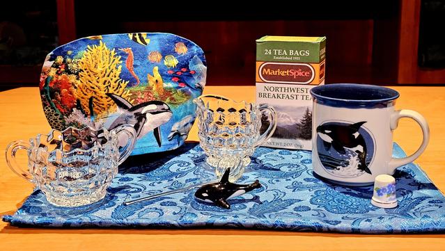 A tea tableau in sympathy with the Orca known as Tahlequah whose newborn calf has died and for which she visibly grieves. On a wavy field of blue is a killer whale figurine surrounded by a handmade mug with an orca design, a cozy cover with an ocean scene including orcas, Depression glass creamer and sugar bowl, a glass sugar spoon shaped like a seashell, a thimble with Forget- Me- Not flowers painted on it, and Northwest Breakfast black tea from Market Spice in Seattle.