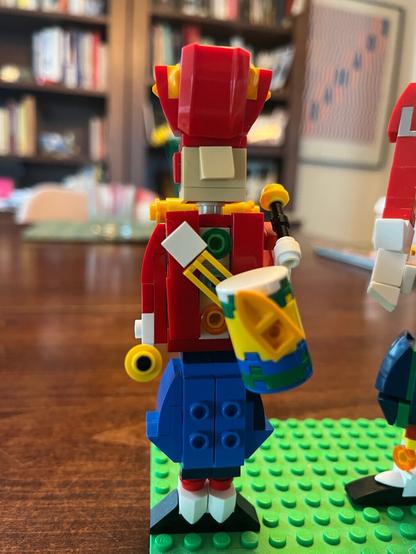 A colorful LEGO solder drummer wearing a red uniform with yellow accents, blue pantaloons, black shoes, white gloves, holding a small drum.
