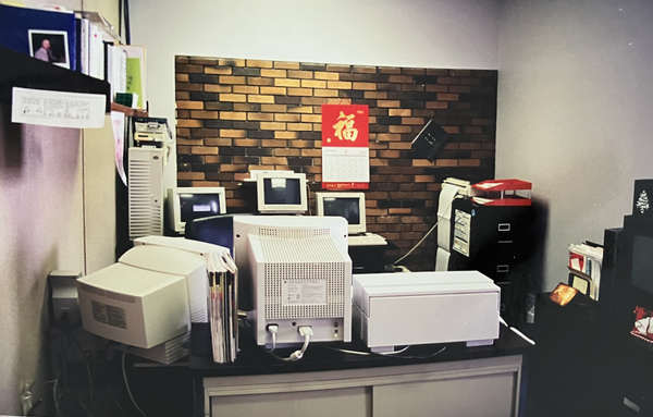 A retro office desk from 1996 with a brick-pattern wall in the background. The desk is crammed with CRT monitors, an IBM tower functioning as a web server, and an ISDN modem stack. Papers, files, and old office equipment complete the scene, capturing a tech-heavy, pre-dot-com-era workspace.