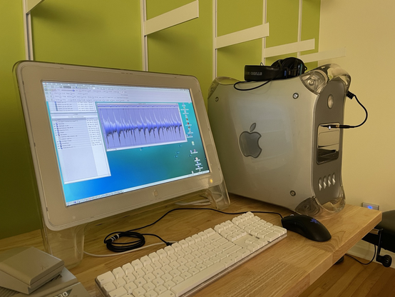 A mac G4 mirrored drive door computer on a bench NEC to to its 20” lucite studio (or cinema?) display. A bright green wall is behind the bench, and there is white shelving hardware on the wall but the shelves have not been installed yet.