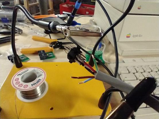 A tiny Mini-DIN connector being soldered to a multi-conductor cable.  The Mini-DIN does not have appropriate terminals on the cable side, it has pins rather than solder cups.  The connector and cable are being held by a helping hands device.  An Apple II GS is visible in the background.