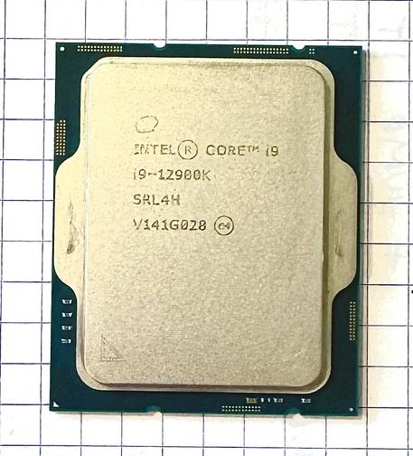 The offender's face. The top side of the Intel Core i9 CPU. Although the PCB of the chip is a lovely deep, dark green, it is mostly obscured by the chip's own heatsink/heat-transfer plate, which is, as the base-board, a portrait-oriented rectangle. It has rounded corners, however, and a stepped bevel, which the PCB does not. It sits in from the edge of the PCB by an almost uniform 3mm. From its left and right midsection, two "ears", of a similar style and curvature, extrude, to bear the force of the LGA socket bracket. Marks from its usage can be seen on these extrusions. The board section has patches of metal dots in little grids here and there, as well as a few top-mounted SMD components. It is sitting neatly aligned on 5mm grid paper. I will spare you the few lines of numbers and letters inscribed on the heatsink surface in four neat lines. A triangle/arrow is also inscribed in the bottom left corner.