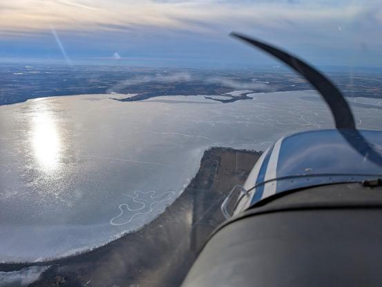 Looking out of front of a Cessna 172 Skyhawk. The blue cowling is visible with the propeller spinning and out the window is a large frozen lake.