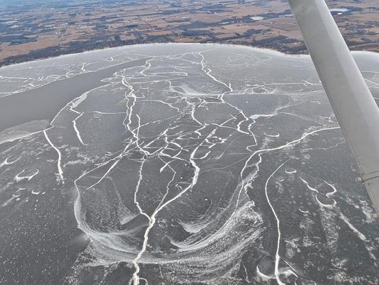 View from above of a lake showing giant fissures in the ice sheet on the lake. There is open water in the top left and some swept snow scattered around showing where it is shielded from the wind due to the ice being non-uniform.