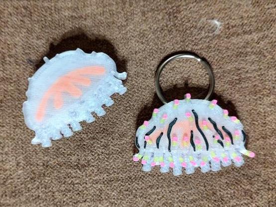 Two 3d printed flower-hat jelly designs. On the right, the "top", showing a translucent jellyfish-shaped creature with an orange inner structure. It has dark colored stripes on the bell, and is covered with many small tentacles that are tipped with green and pink. It has a 1-inch (2.5cm) spit ring through the top. The one on the left is upside down, showing only the clear body and the orange inner structure.