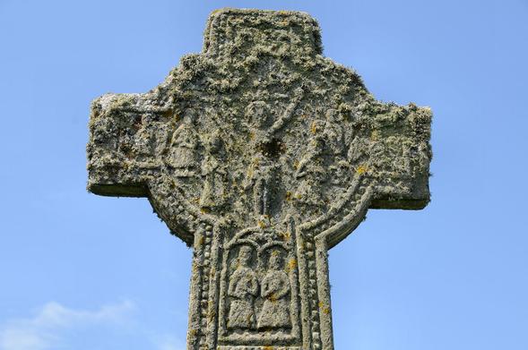 The head of the beautiful 2.57m high Kilchoman Cross. The image is looking up at the head of the cross, which has a large circular area with depictions of figures and more figures carved below at the top of the shaft. The arms of the cross are quite short The stone of the cross is heavily overgrown with lichen. The sky is blue.