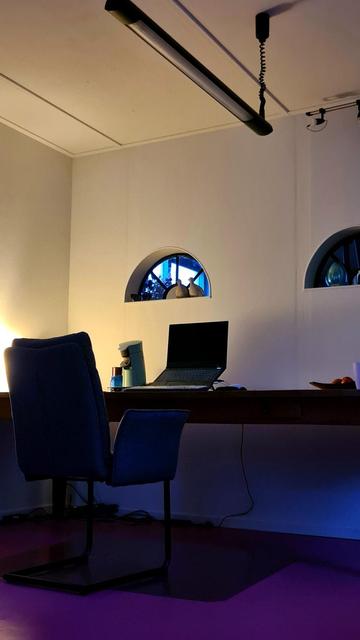 A photo of a workspace. A comfortable chair at a long desk with a laptop on a stand and a coffee maker next to it. The room has a clean modern look to it, the floor is bright pink and the wall is white and has two spoked semi circular windows with small statues standing in them.