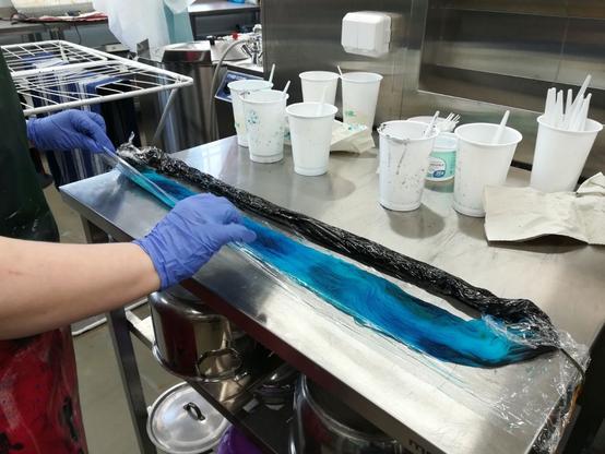 Työtilassa Veera rullaa vyyhdin kelmuun rullalle kuumennusta varten. Vyyhdin takana valkoisissa muovipurkeissa on värinesteet ja muita härpäkkeitä.