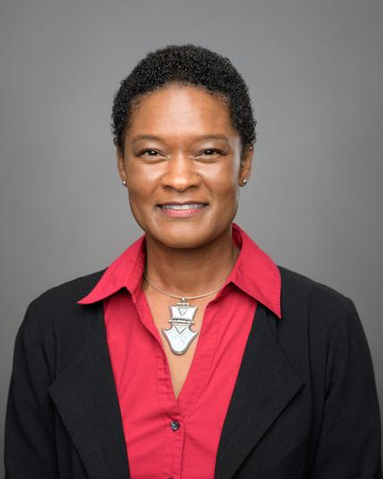 Tracy Drain, a Black woman with short hair, wearing a black blazer over a red top and a large silver pendant necklace, smiling in front of a grey backdrop.