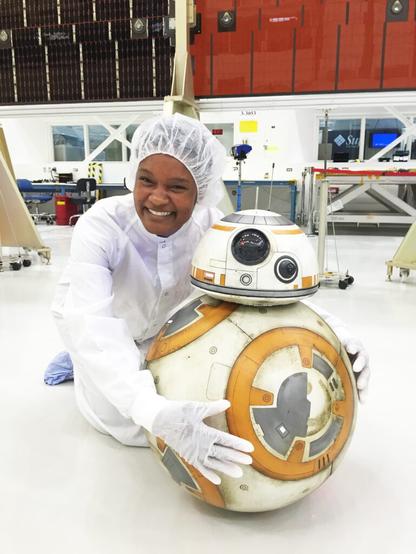 Tracy Drain, a Black woman wearing full-body white clean suit, kneeling on the floor of a technology lab, smiling broadly and hugging BB-8, the spherical orange-and white Star Wars droid.