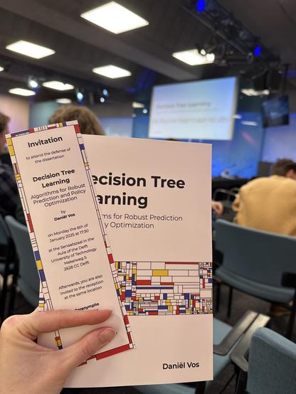 View from the audience of a conference room. I am holding up a dissertation and bookmark. Their designs are clearly inspired by the work of Piet Mondrian.
The bookmark contains the following text:
Invitation
to attend the defense of the dissertation
Decision Tree Learning
Algorithms for Robust Prediction and Policy Optimization
by
Daniël Vos
on Monday the 6th of
January 2025 at 17:30
at the Senaatszaal in the
Aula of the Delft
University of Technology
Mekelweg 5
2628 CC Delft
Afterwards, you are also
invited to the reception
at the same location
